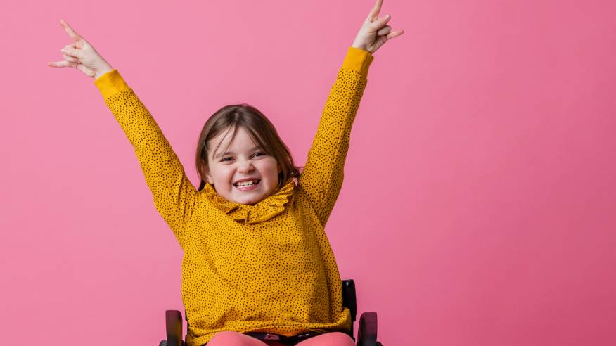 A girl wearing a yellow jumper sits in a wheelchair. She holds her hands in the air as she grins.